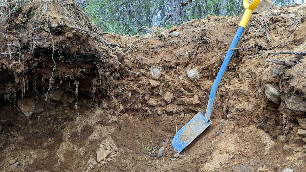 Eine Schaufel steht in einer Grube, die ein Goldgräber in Tankavaara ausgehoben hat. Das Foto zeigt das alte Flussbett im Querschnitt.  