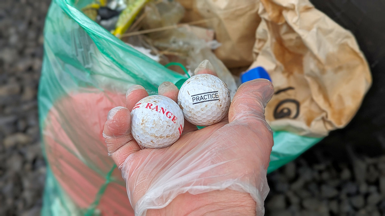 Eine Hand hält zwei gefundene Golfbälle vor einer Plastiktüte mit aufgesammelten Abfall