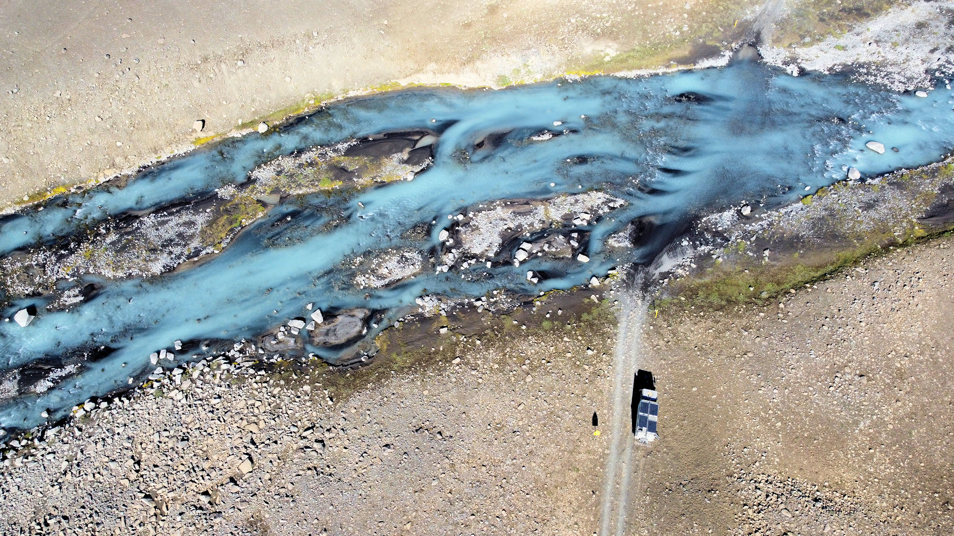 Luftbild von einer Furt in einer steinigen Landschaft ohne Vegetation. Der Fluss hat milchiges Wasser. Unten wartet ein LKW darauf, durch die Furt zu fahren. 