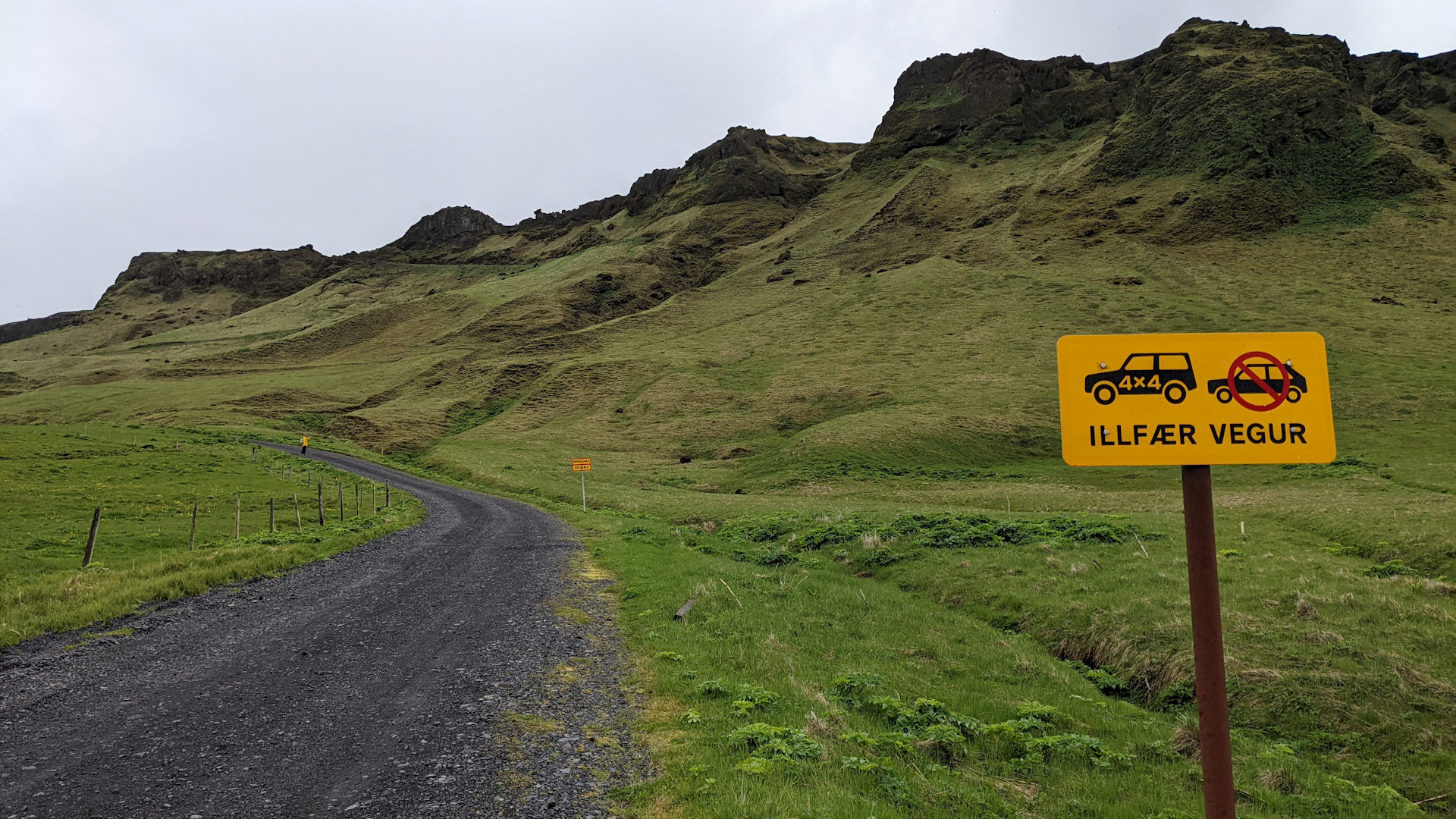 Eine Schotterpiste führt in grüne Berge hinein. Rechts steht ein Schild, auf dem ein 4x4 Geländewagen abgebildet ist und ein gewöhnliches Auto. Das gewöhnliche Auto ohne 4x4 Antrieb ist rot durchgestrichen.  