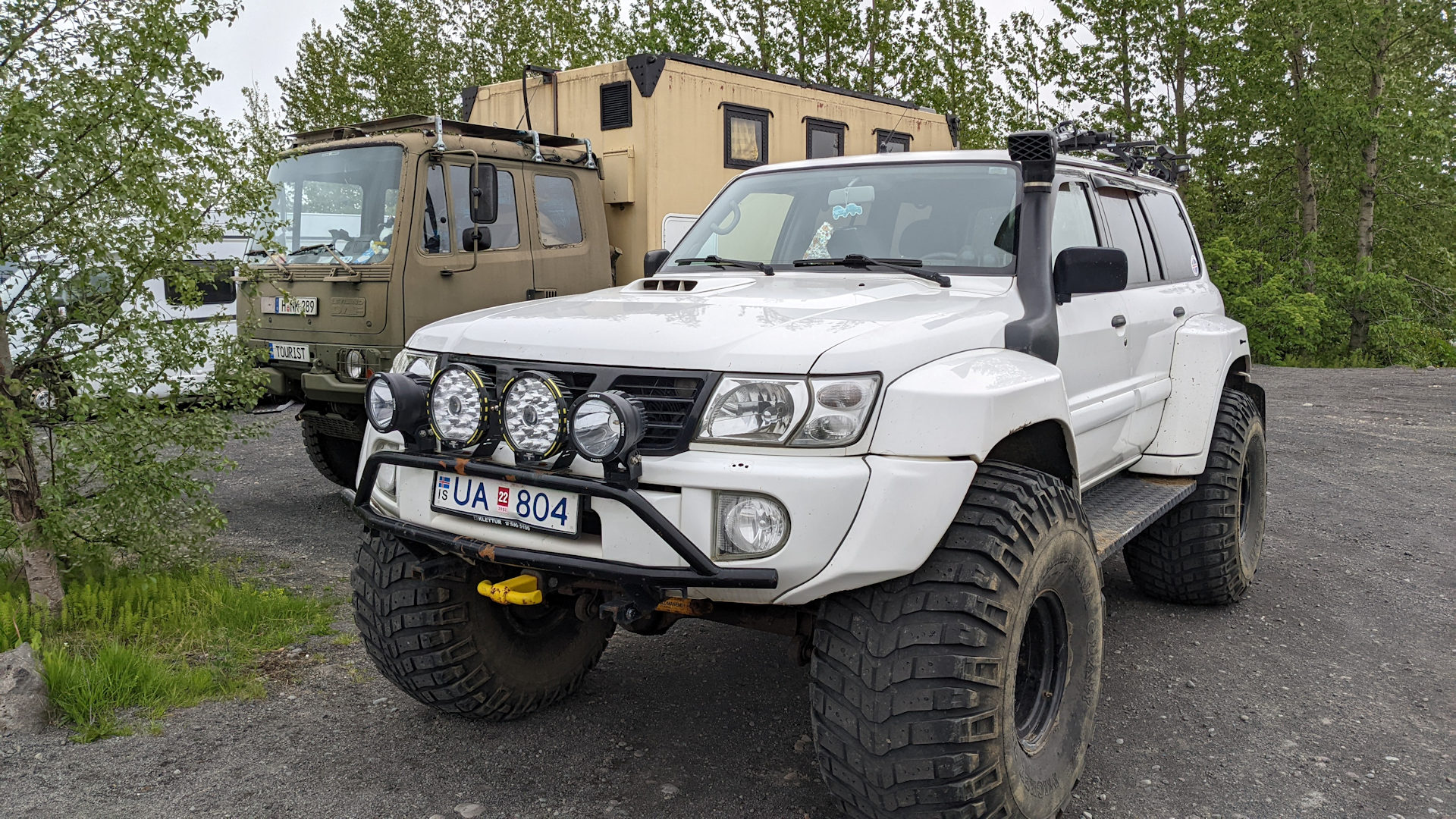 Ein weisser Geländewagen mit extra breiten Reifen und zusätzlichen Lampen und einem gelben Haken zur Bergung steht vor einem halb verdeckten DAF T244 Gelände LKW. 