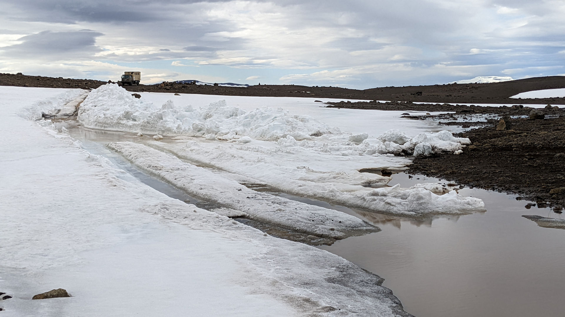 Ein DAF T244 LKW wartet im Hintergrund vom Bild darauf, von oben links nach unten rechts über eine mit Schnee bedeckte Piste zu fahren. Die Piste führt durch ein Schneefeld und endet unten rechts in einer Wasser Lache