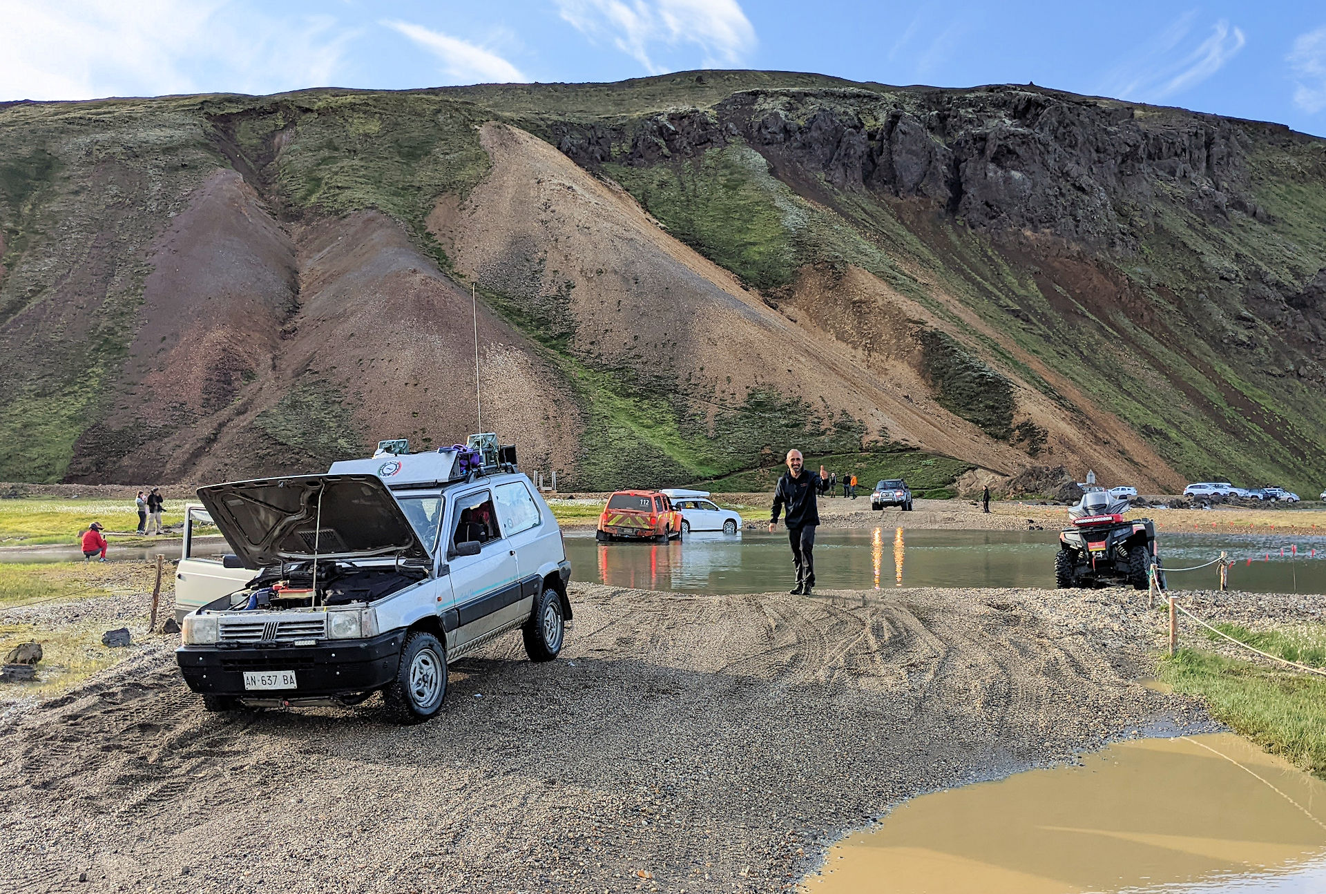 Links steht ein kleiner Fiat 4x4 Geländewagen mit geöffneter Motorhaube. Im Hintergrund sind eine wassergefüllte Furt, Berge und ein paar andere Fahrzeuge zu sehen.  