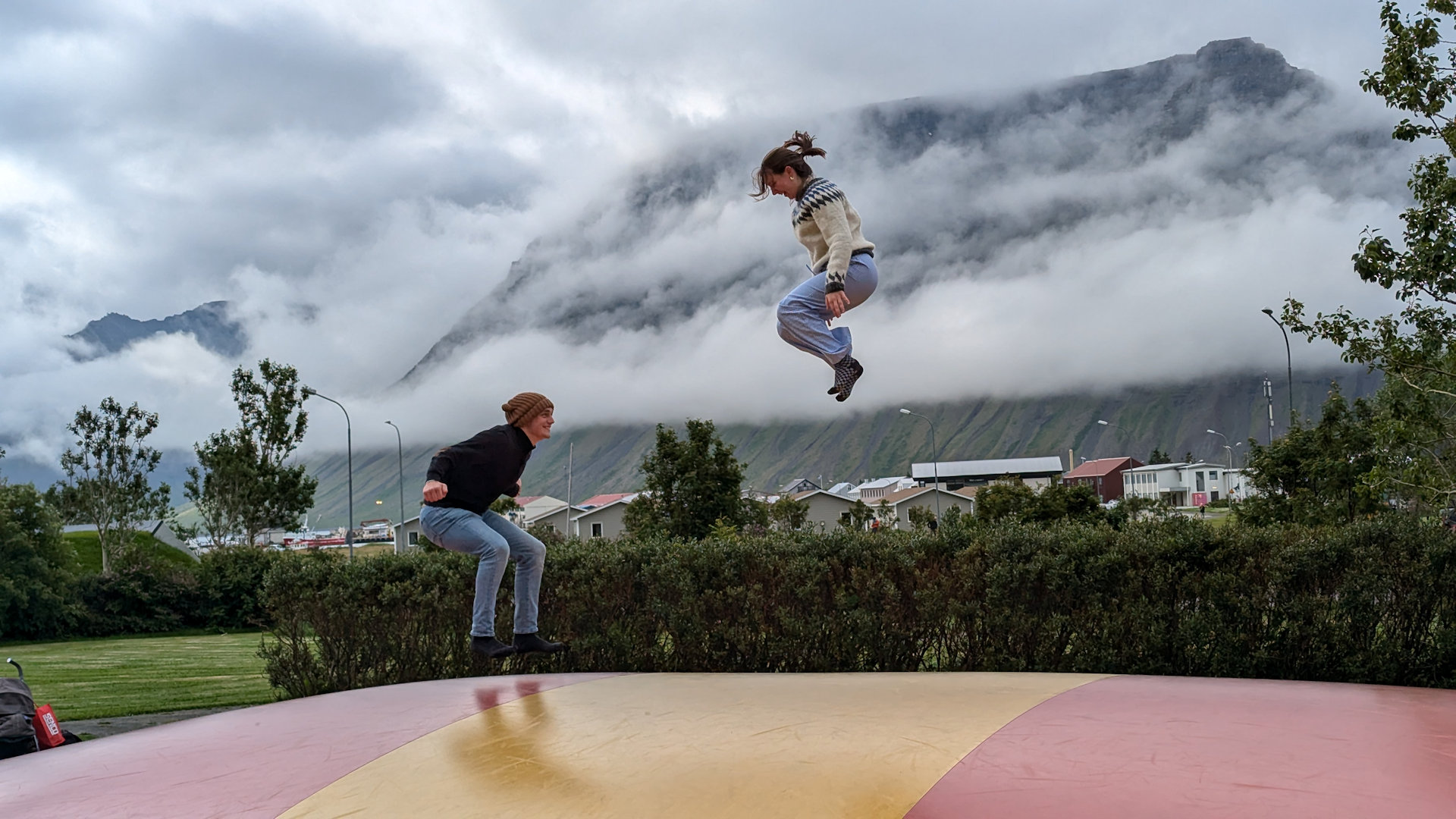 Zwei Jugendliche auf einem Trampolin vor wolkenverhangenen Bergen in Island