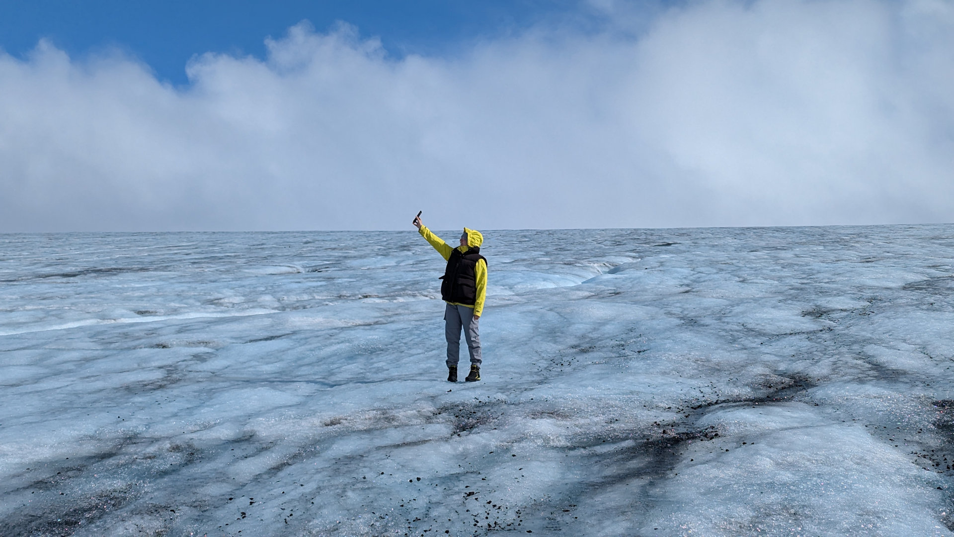 June Frau in gelben Regencape hält auf einem Gletscher ein Handy für ein Selfie in die Höhe