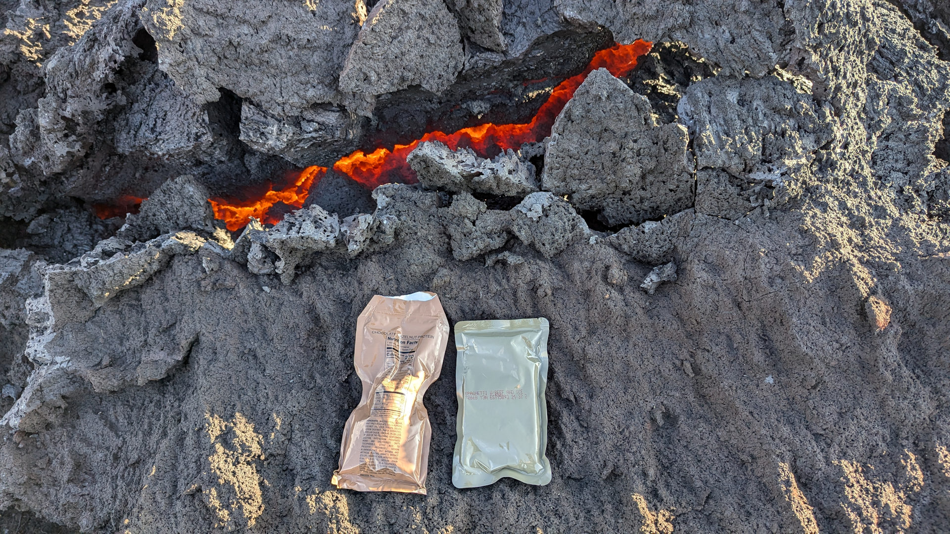 Zwei MRE Mahlzeiten werden auf heissen Steinen erwärmt, in Hintergrund ist rot glühende Lava