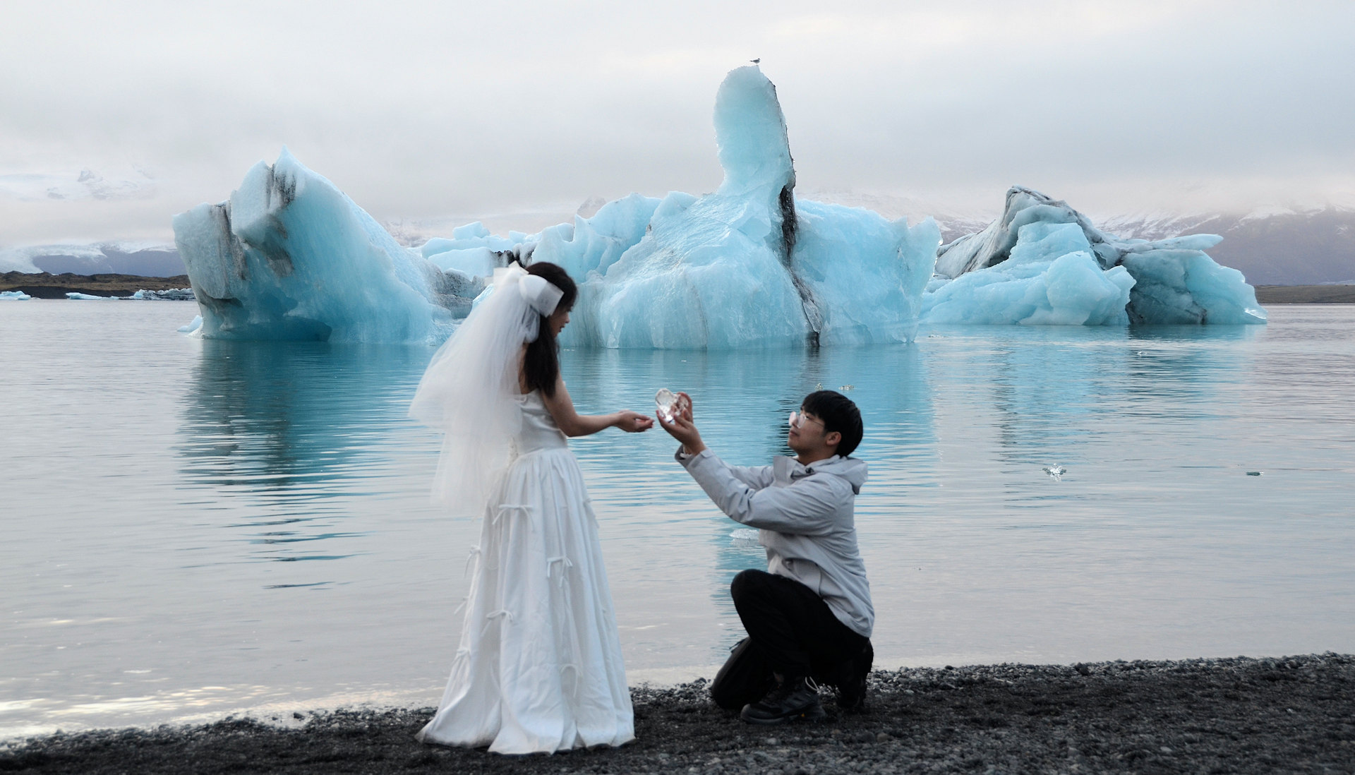 Einer chinesischen Braut wird am Jökulsarlon vom knienden Bräutigam ein Herz aus Eis überreicht. Blau schimmerndes Eis im Hintergrund.  