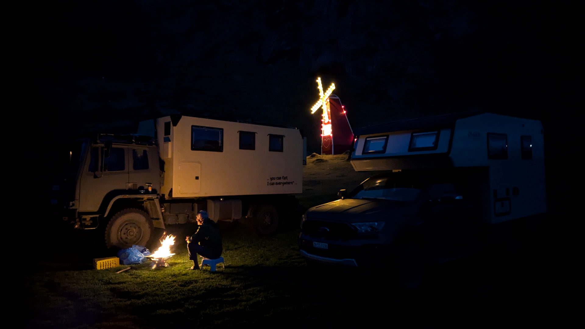 Mann sitz an einem Lagerfeuer zwischen einem LKW und einem dunklen Geländewagen 